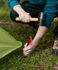 Person using a mallet to drive Ultralight 7-Inch Aluminum Tent Stakes Set 12 Pack With Reflective Pull Ropes into the ground