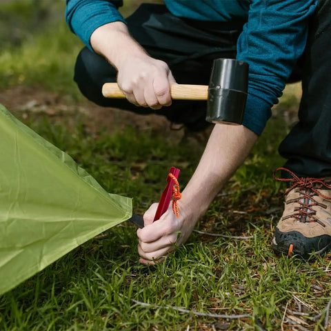 Person using a mallet to drive Ultralight 7-Inch Aluminum Tent Stakes Set 12 Pack With Reflective Pull Ropes into the ground