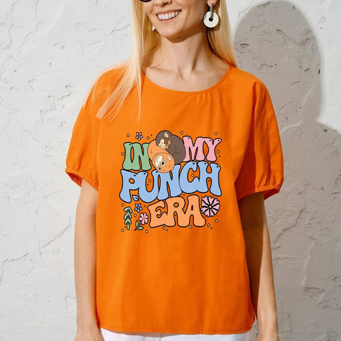 Woman in orange shirt with 'In My Punch Era' graphic, showcasing color text and cartoon bears with floral accents and sunglasses.