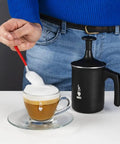 Tutto Crema Milk Frother Black Barista Grade, 1L Capacity with double mesh, next to a frothy coffee cup on a table.