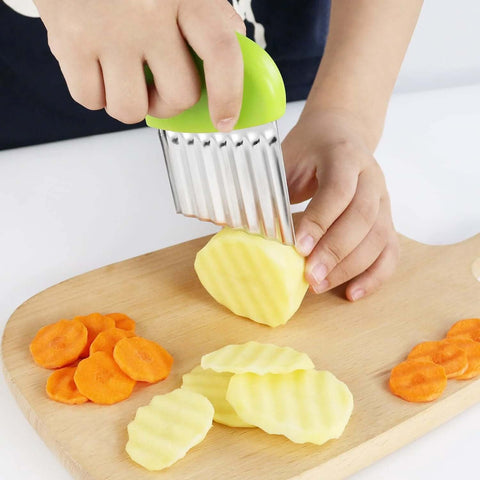 Two stainless steel crinkle cutters with green handles, slicing potatoes and carrots into wavy chips on a board.