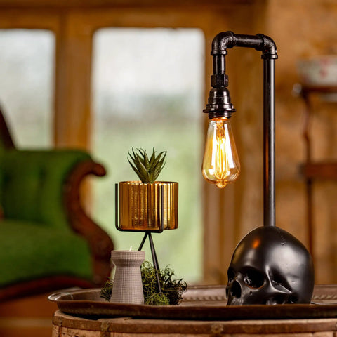 Black Industrial Skull Lamp With Skeleton Decor For Gothic Bedroom Dope Handcrafted Cast Iron And Resin Skull Table Lamp Vintage Industrial Vibe UL Certified 18.5 Inches Tall 1 to 40W Bulb Not Included on wooden table with plant; rustic background