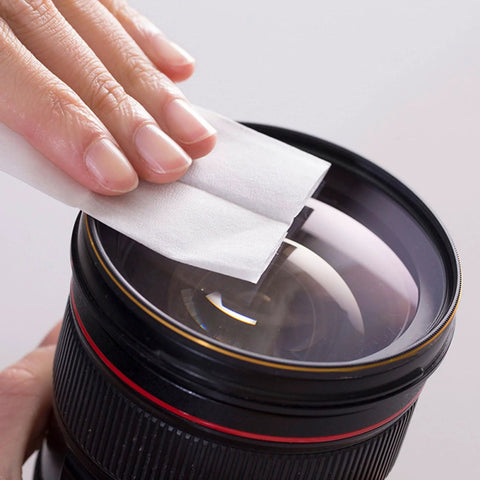 Lens cleaning paper tissue being applied to a camera lens; white 10x15 cm wipe held by fingers