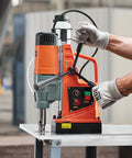 Magnetic drill press on metal worktable, orange and gray body, operator wearing gloves using the feed handle.