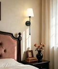 KSANA Modern Black Wall Scone Lighting Indoor with White Fabric Shade next to a bed with decorative elements.