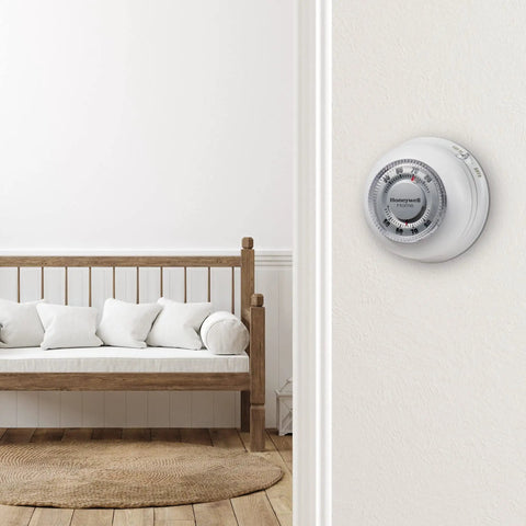 Round No Frills Non Programmable Manual Thermostat with Easy Dial Control in a cozy home setting, wall-mounted near a wooden bench.
