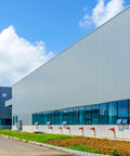 Industrial warehouse side view with gray metal siding, blue-tinted windows, and red pipes along the wall.