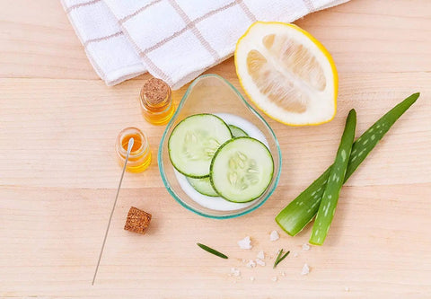 Sliced cucumbers in a bowl with aloe vera, lemon half, essential oils, and a towel on a wooden surface, spa ingredients.
