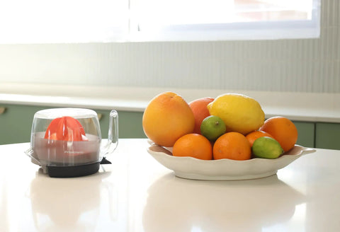 Primtronic Electric Citrus Juicer With Large 24 Oz Capacity And Seed Filter Auto Reverse Function Compact Design For Oranges Lemons Limes BPA Free Black Orange next to a bowl of fresh citrus fruits.