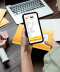 Person holding a phone with kraft bubble mailers in background, Kraft Bubble Mailers 4x8 Inches 50 Pack Self Adhesive Padded Envelopes visible