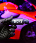 Close-up of gray GYEON MOHS ceramic coating bottle held by gloved hand, automotive detailing product with neon car.