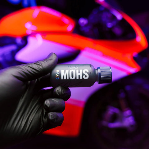 Close-up of gray GYEON MOHS ceramic coating bottle held by gloved hand, automotive detailing product with neon car.