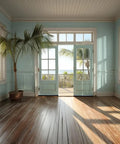 Sunlit room with wooden floor, blue walls, potted palm, and glass doors overlooking a balcony with ocean view.