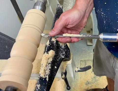 Carbide wood turning tool with 12-inch aluminum handle guiding on a wood lathe, beige turned workpiece.