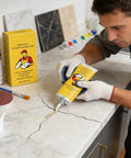 Man using Fast Dry Tile Repair Glue Kit For Cracks Chips And Gaps On Ceramic Porcelain Floor And Wall Tile Surfaces Waterproof Quick Set Adhesive Beginner Friendly DIY Fix For Stone And Tile Cracks on a cracked marble countertop, with yellow packaging and pigment set visible in the background.