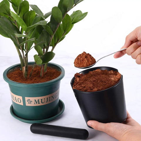 Hand using spoon to transfer coffee grounds from black container near plant, removable knock bar visible. Espresso Knock Box And Tamper Mat Set For Baristas With Removable Knock Bar Non Slip Base Food Safe Silicone Grounds Disposal Easy Cleanup Great For Home Cafe Or Busy Coffee Shop Perfect Gift For Enthusiasts And Pros.