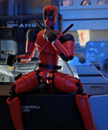 Red and black action figure sitting on a table with blue background, near a Bambu Lab AMS 3D printer.