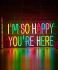 Colorful LED sign with text "I'm So Happy You're Here" on brick wall, vibrant neon lights reflecting on table surface.