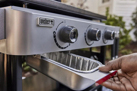 Hand removing drip tray from stainless steel grill with control knobs visible, part of Griddle Essential 5 Piece Tool Set.