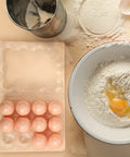 Clear Plastic Egg Cartons Bulk 200 Pack 3x4 Grid Stackable Trays For Farm Market And Home Use See Through Design Keeps Eggs Secure And Easy To See In Fridge with eggs next to a mixing bowl and flour sifter.