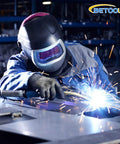 Betooll welder in blue coveralls with helmet welding metal plate; sparks fly.