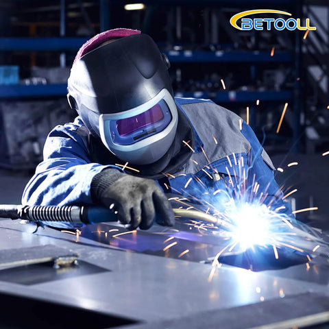 Betooll welder in blue coveralls with helmet welding metal plate; sparks fly.