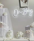 Oh Baby neon sign in cool white above white draped table with floral decor, part of Lit Neon Sign For Wall Decor 31.5 By 15.7 Inches With Dimmable Switch Reusable Neon Light Sign For Baby Shower Birthday Party And Wedding Decor In Cool White.
