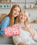 Two women smiling, one holding a pink gift box labeled 'Best Wishes' in a cozy home setting.