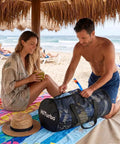 Couple at beach with 100L Duffel Scuba Bag For Diving And Watersports, Large Mesh Gear Tote with towels on sand.