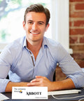 Man smiling at desk with computer, wearing blue shirt, sits next to business card that says 'ABBOTT, The company directooo'. Bright background.