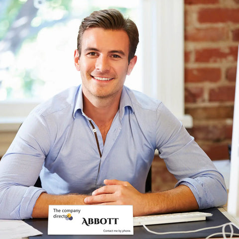 Man smiling at desk with computer, wearing blue shirt, sits next to business card that says 'ABBOTT, The company directooo'. Bright background.