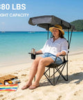 Person sitting on oversized camping chair with adjustable canopy on a beach. Chair has 380 lbs capacity and padded armrests.