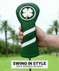 Hand holding a green leather golf club head cover with clover emblem on a golf course background.
