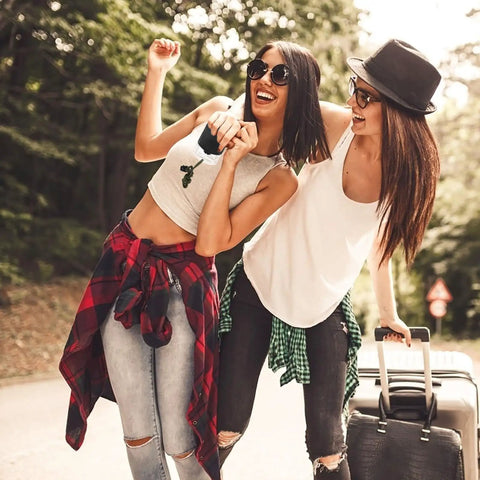 Two people smiling and holding luggage outdoors in casual clothing, enjoying the sunny day