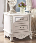 White ornate bedside table with floral carvings and two drawers featuring vintage leaf drawer pulls in a rustic setting.