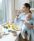 Mother holding baby while drinking water in modern kitchen with healthy breakfast items on the counter.