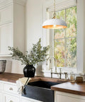 Top Shelf Matte White Farmhouse Pendant Light With Burnished Brass Accents above a kitchen sink with a dark stone counter.