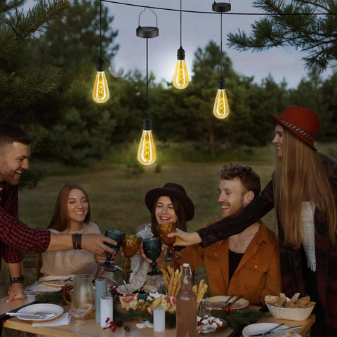 Two Pack Hanging Solar Lights With Glass Edison Bulbs Solar Powered Lanterns Outdoor Waterproof LED Garden Decor For Patio Yard And Tree Cozy Candlelight Ambience For Parties And Relaxed Evenings over dining table with people toasting outdoors.