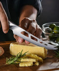 Five Inch Serrated Cheese Knife Crafted With Premium Steel Core And A Super Slick Hollow Blade For Butter Smooth Cuts That Slide Off Easy Plus A Gotta Have Fork Tip For Zero Mess Party Spread, slicing cheese on a wooden board.