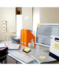 Pop-up sticky note dispenser with one 50-sheet canary yellow 3 in x 3 in pad, black base and clear lid, on office desk setup