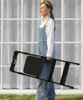 Lightweight Aluminum Three Step Folding Ladder With Big Wide 13.8 By 13 Inch Platform And Handy Tool Tray For Home Projects, ANSI A14.2 Certified, Sturdy Portable Stepladder For Indoor Fixes And DIY Tasks. Woman carrying a folded black aluminum 3-step ladder with wide platform.