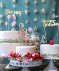 Decorated cakes with gold and pink birthday toppers, featuring 'Hooray,' '23,' and 'Happy Birthday' in festive display.