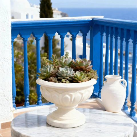 Succulent arrangement in an ornate white planter on a marble table, overlooking a coastal view with blue railings and vase.