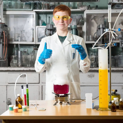 Young scientist in lab coat and safety goggles giving thumbs up, conducting experiment with beakers and test tubes on desk.