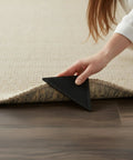 Hand placing Heavy Duty Rug Gripper Pads With Adhesive Strips For Non Slip Carpet Corners Kit Includes Four Pads And Four Auxiliary Stickers Edge Guard And Extra Grip For Home Or Office under a carpet edge on wooden floor