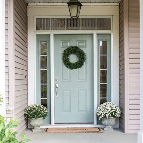 Green wreath on blue front door with side windows, flanked by potted white flowers on a porch
