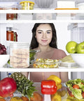 Woman standing behind an open fridge filled with fruits and vegetables, with 20 Pack 4oz Clear Plastic Jars With Lids on the shelf.