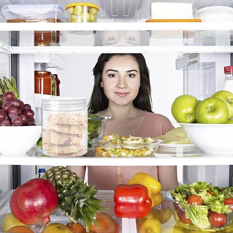 Woman standing behind an open fridge filled with fruits and vegetables, with 20 Pack 4oz Clear Plastic Jars With Lids on the shelf.