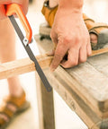 Close-up of hacksaw set with 12 inch steel frame replaceable blades and metal miter guide cutting a piece of wood on a workbench.