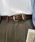 Person wearing a brown belt with a gold metal buckle over gray pants and a white shirt